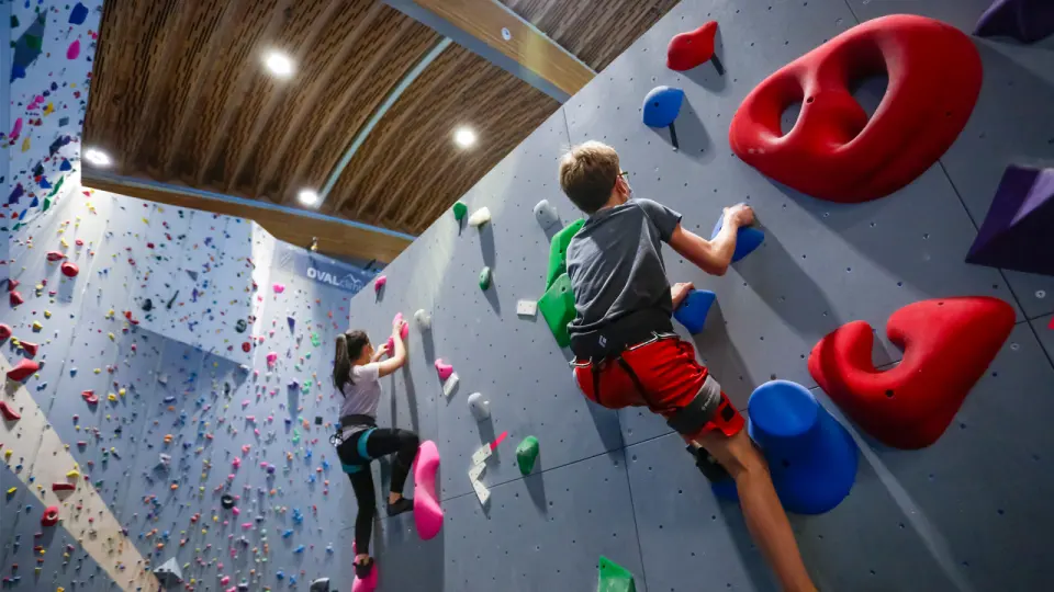Two young climbers on the grey wall below the Oval's curved wooden roof, with the tall rope wall behind