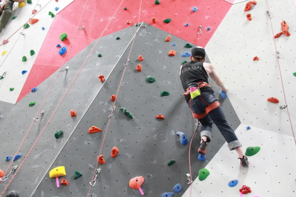 Climber on a grey panel below the red section of the roped wall