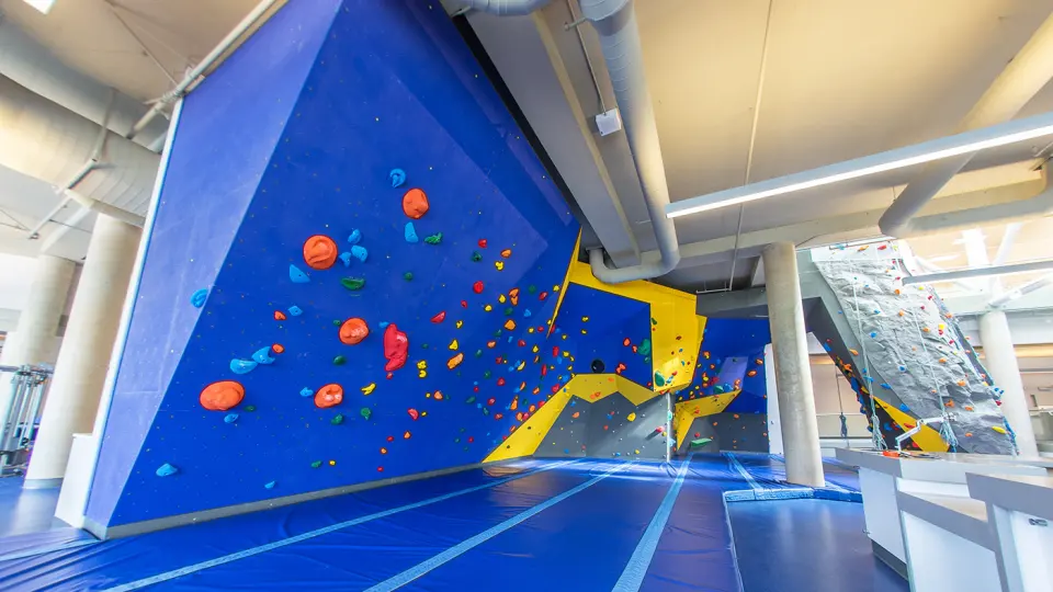 Blue and yellow bouldering wall above blue matting, with the climbing tower beyond