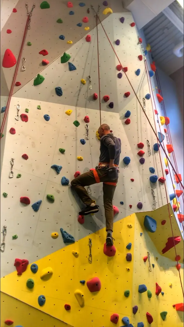 Climber on a tall roped wall with grey and yellow panels and colourful holds