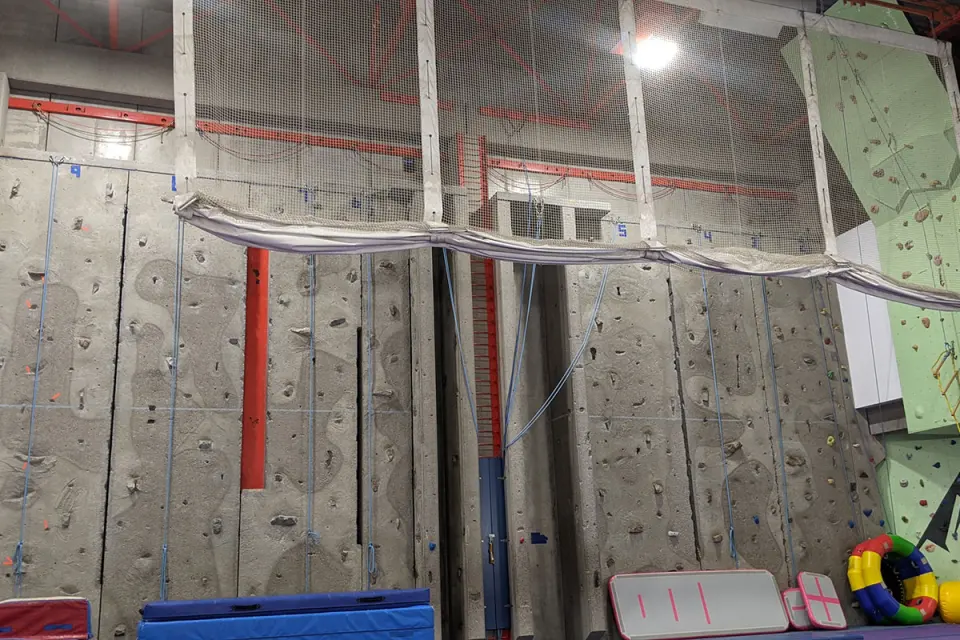 Concrete-textured top-rope wall with blue ropes, safety netting above and mats at its base