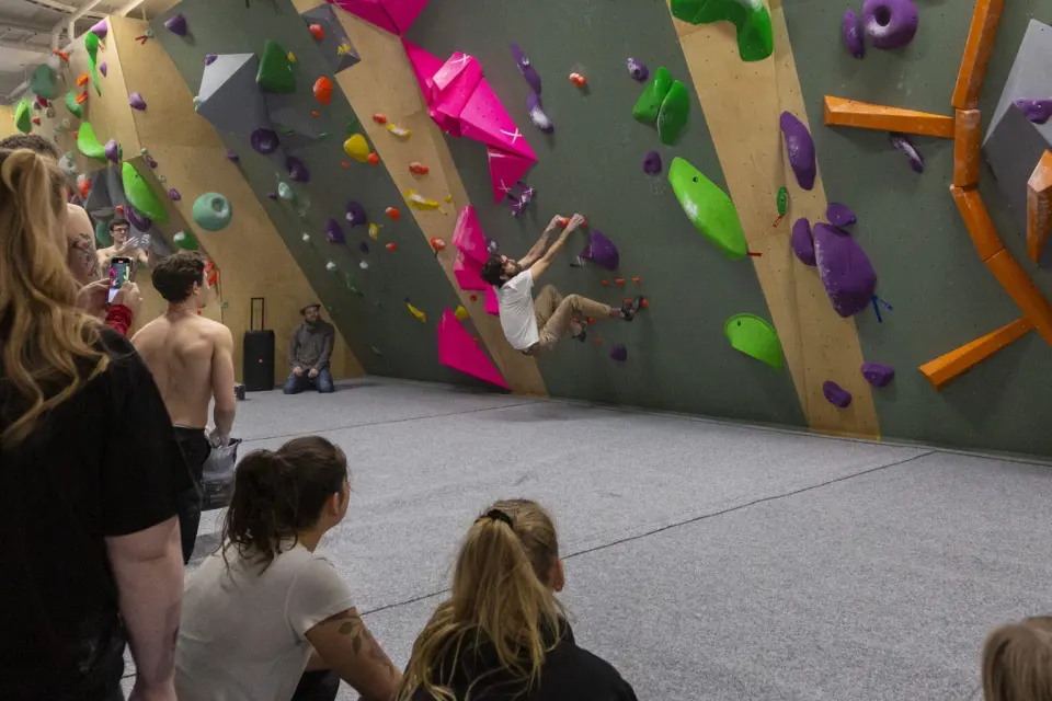 Climber on a steep green bouldering wall with pink volumes, watched by a crowd