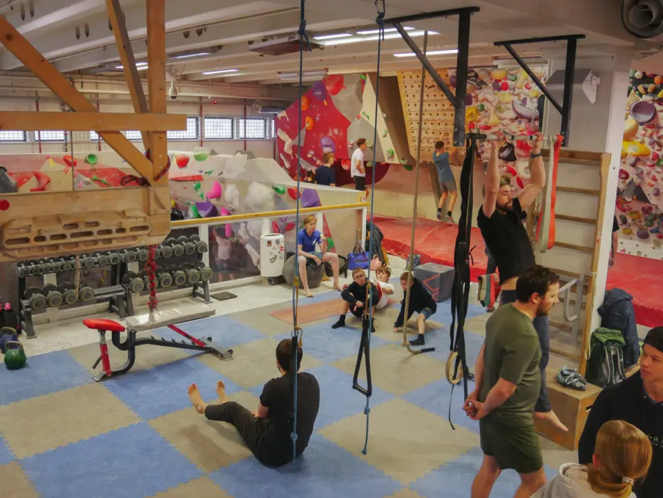 Upper-floor training area with dumbbells, rings and hangboards in front of the bouldering walls
