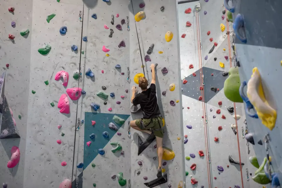 Climber on a tall grey rope wall with coloured holds and volumes