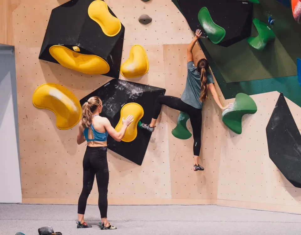 Two climbers on a plywood bouldering wall with black and yellow volumes