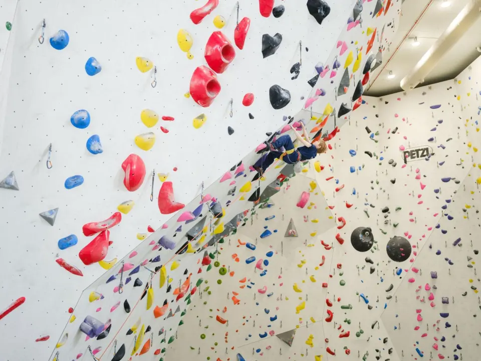 Climber leading up a tall white wall packed with colourful holds and volumes