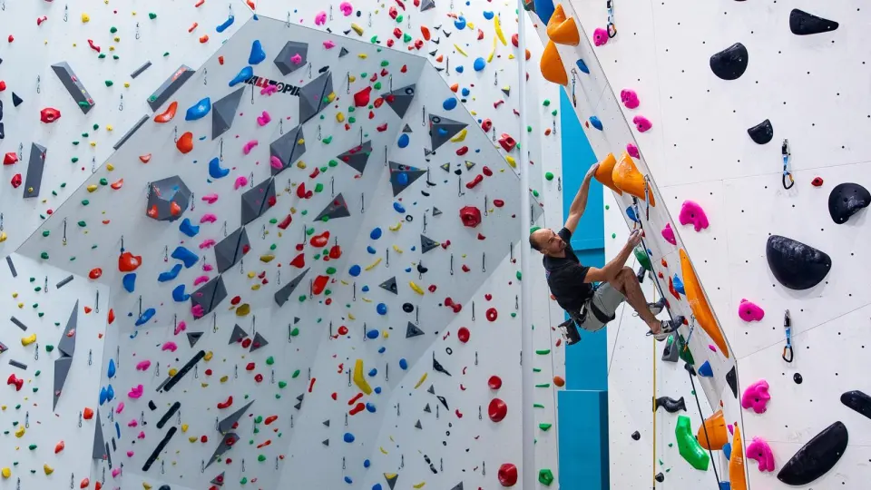 Climber on an overhanging lead route beside a tall white wall of colourful holds