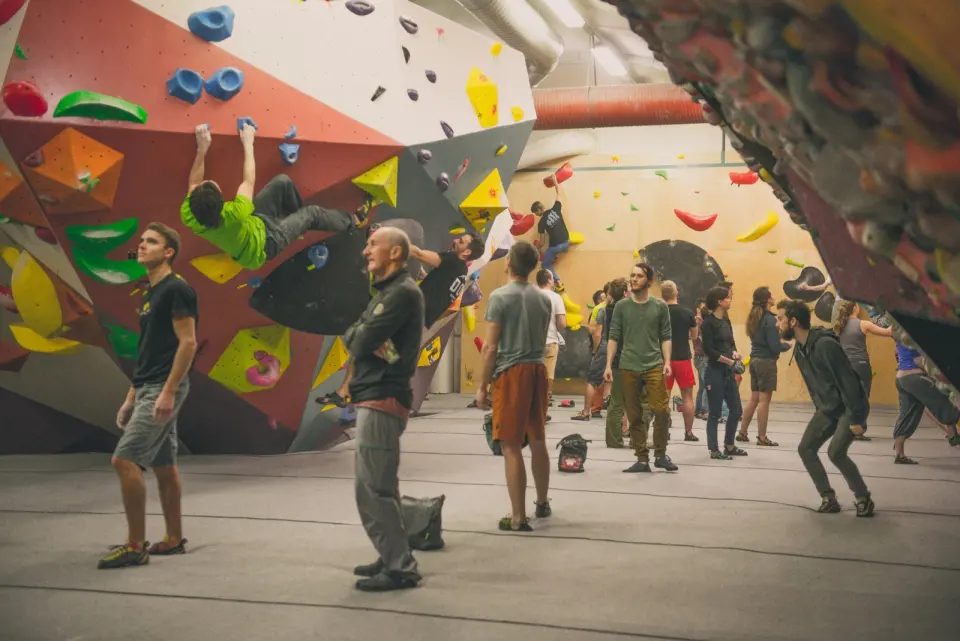 Boulderers on red and white angled walls with big volumes, others watching from the mats