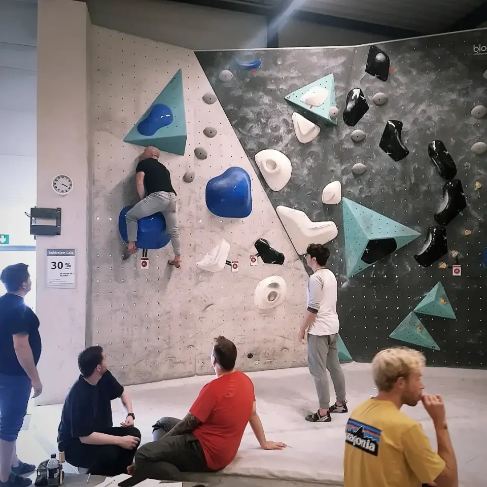 Bouldering wall with blue, white and black holds and teal volumes, climbers resting on the mats