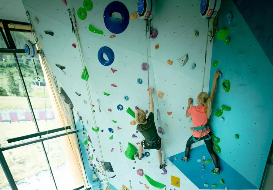 Climbers on auto-belay routes up a tall blue and white wall beside large windows