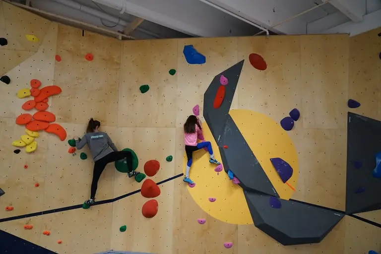 Two climbers on a plywood bouldering wall with a large yellow circle and dark grey volumes