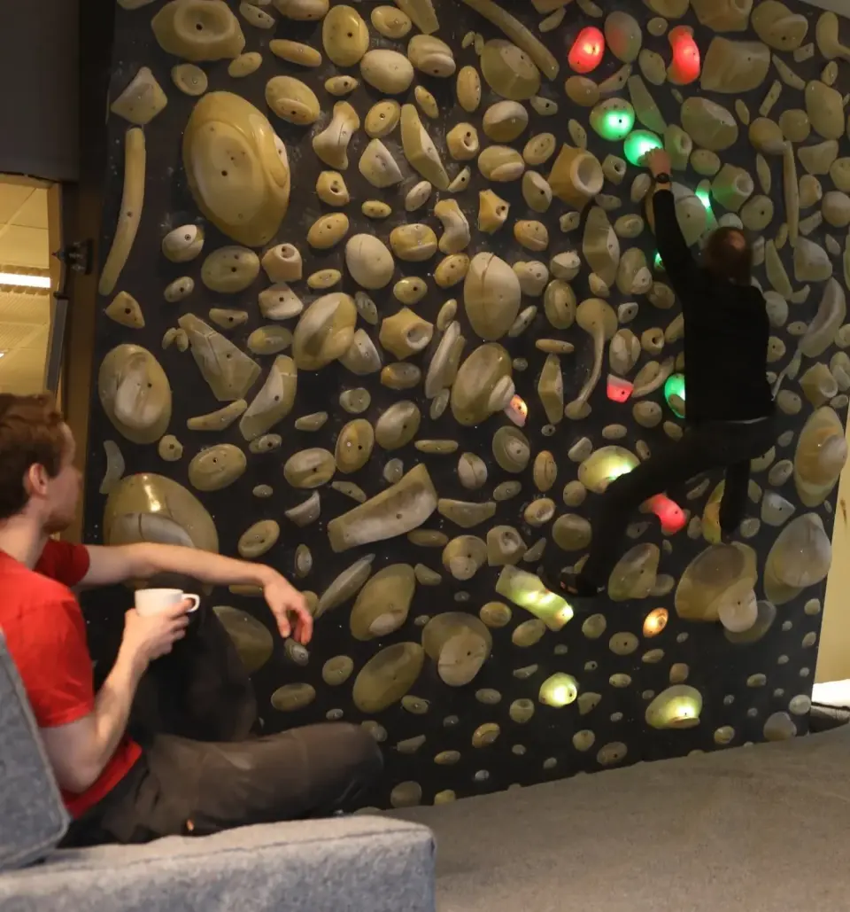 Climber on a dark board wall packed with holds, some lit green and red, while a person watches from a sofa