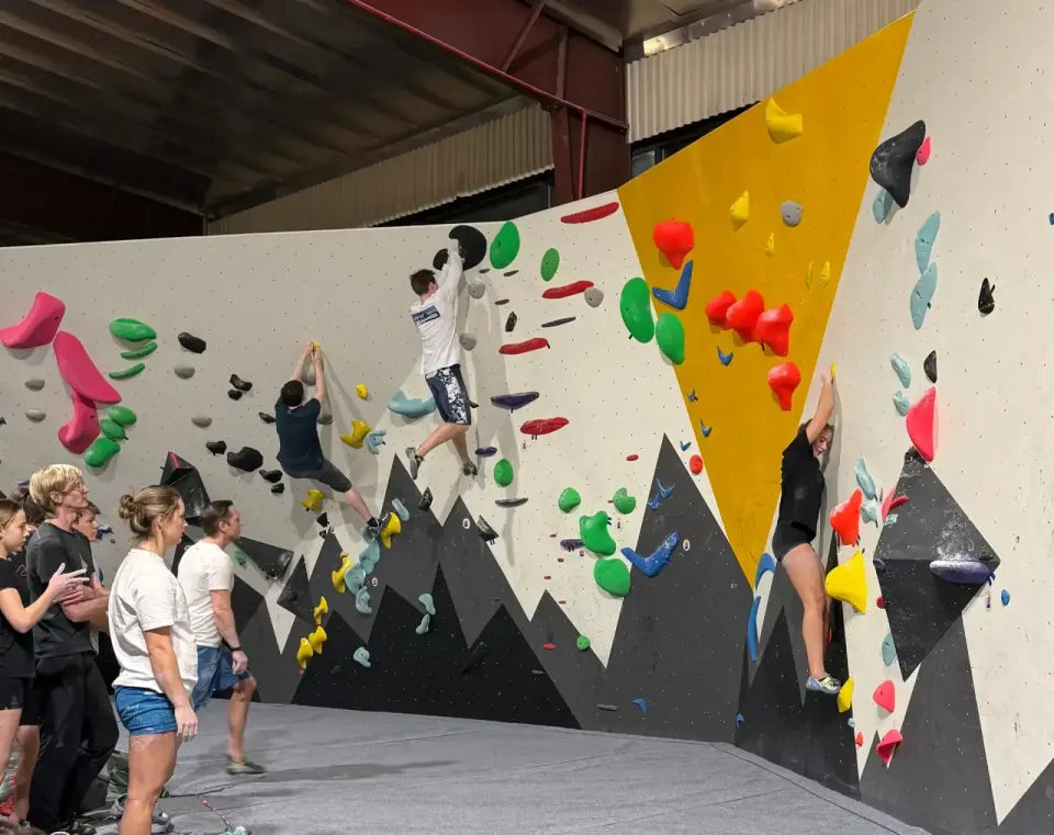 Climbers on a long white bouldering wall with yellow and black panels while others watch from the mats
