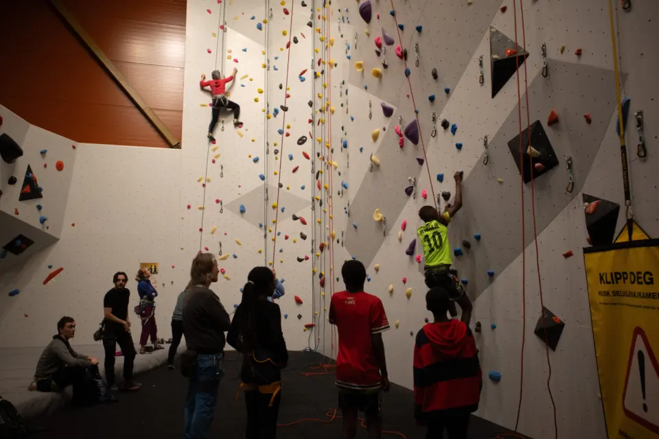 Climbers on top ropes on a tall white wall, with belayers and others waiting on the floor