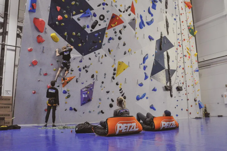 Climber leading up a tall wall of coloured holds and volumes while others belay and rest on the mats