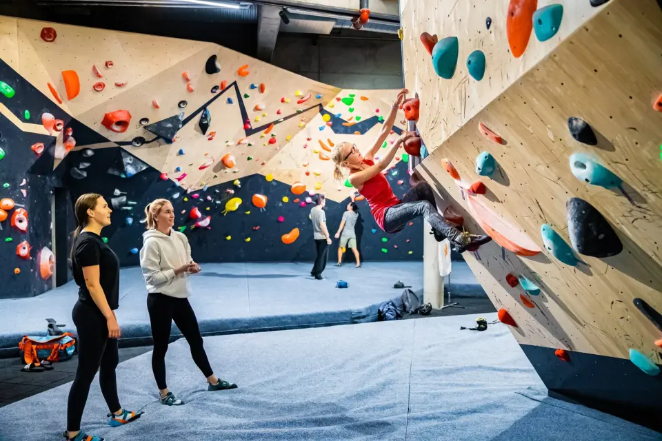 Climber on an overhanging boulder while others watch on the padded floor of the bouldering hall