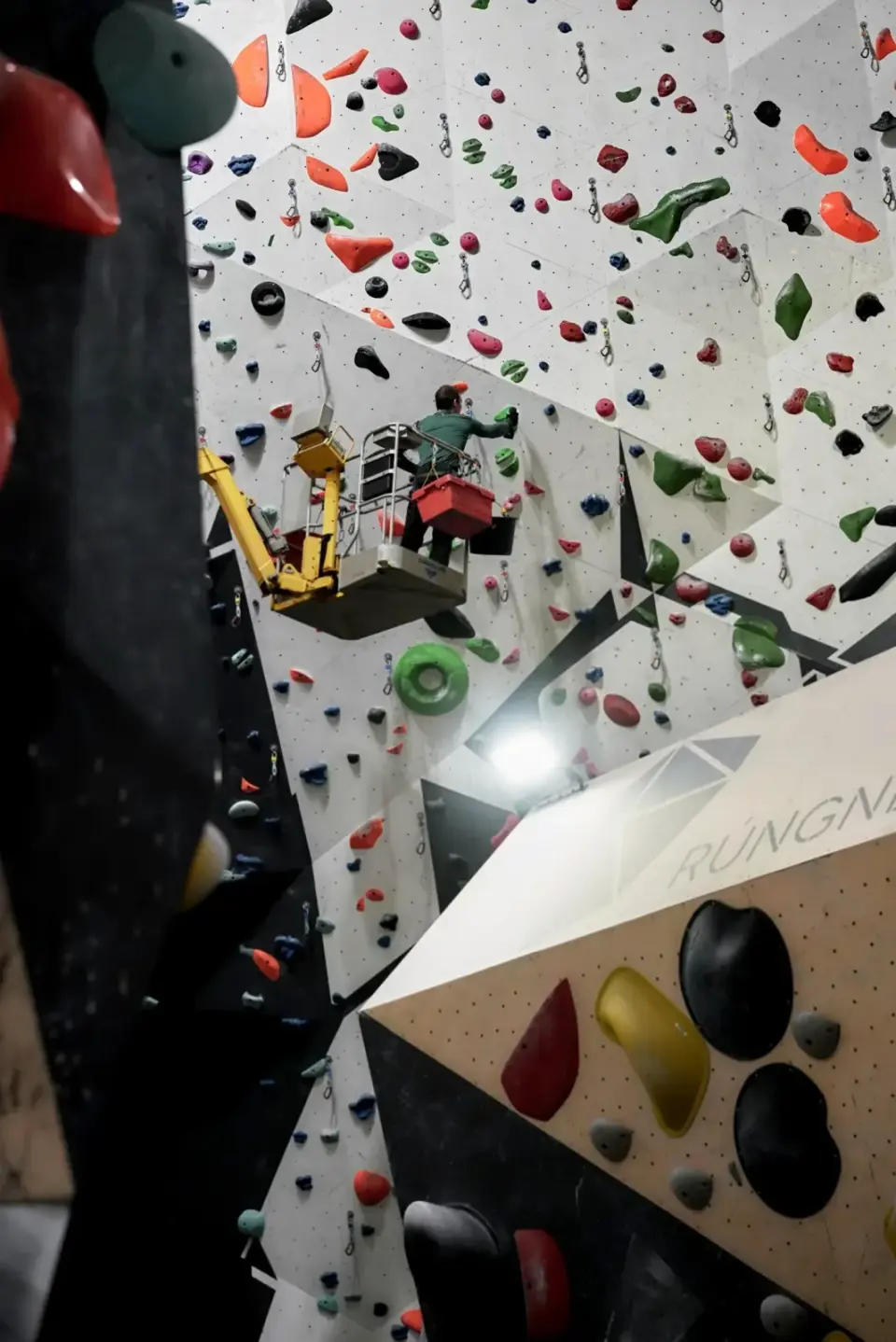 Tall rope wall with a route setter on a lift, seen past a boulder