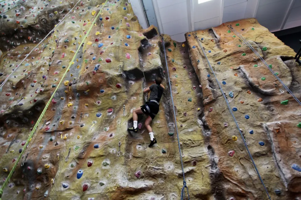 Climber halfway up a tall rope wall with a textured, rock-like surface, seen from above