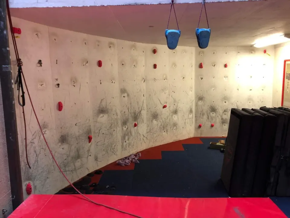 Curved bouldering wall with red holds over carpeted floor, with hanging grips and stacked crash pads