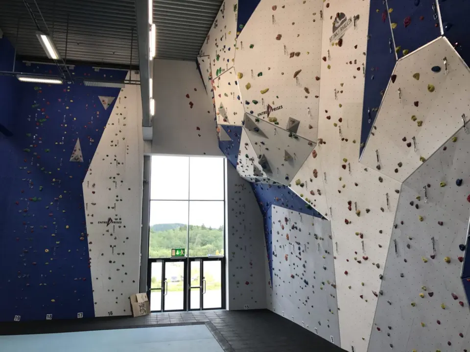 Tall white and blue climbing walls with numbered rope lines and quickdraws in a sports hall