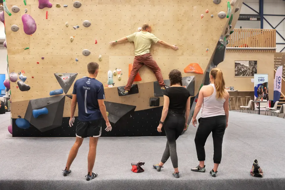Coach and climbers watching a boulderer on a plywood wall, with café seating and a loft behind