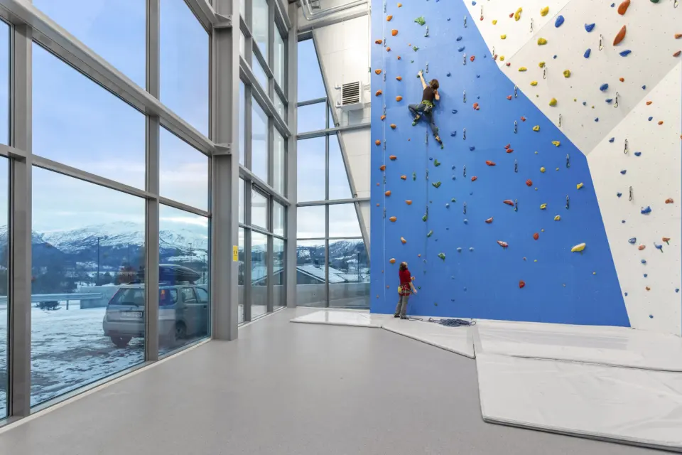 Tall blue and white rope wall beside floor-to-ceiling windows, with a climber and a belayer