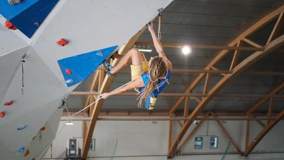 Lead climber clipping a quickdraw on a grey overhang beneath the hall's arched wooden roof