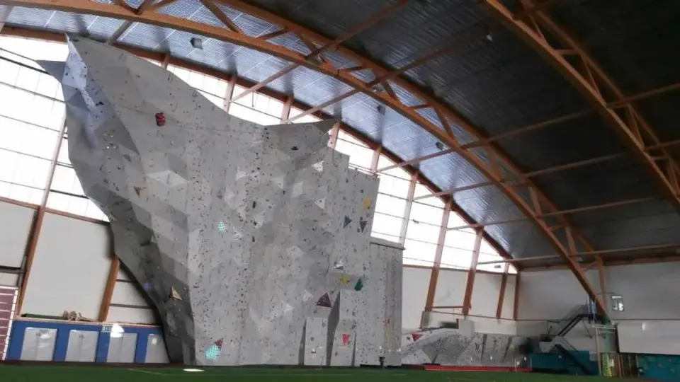 The tall grey main climbing wall with a big overhang at the end of an indoor football hall