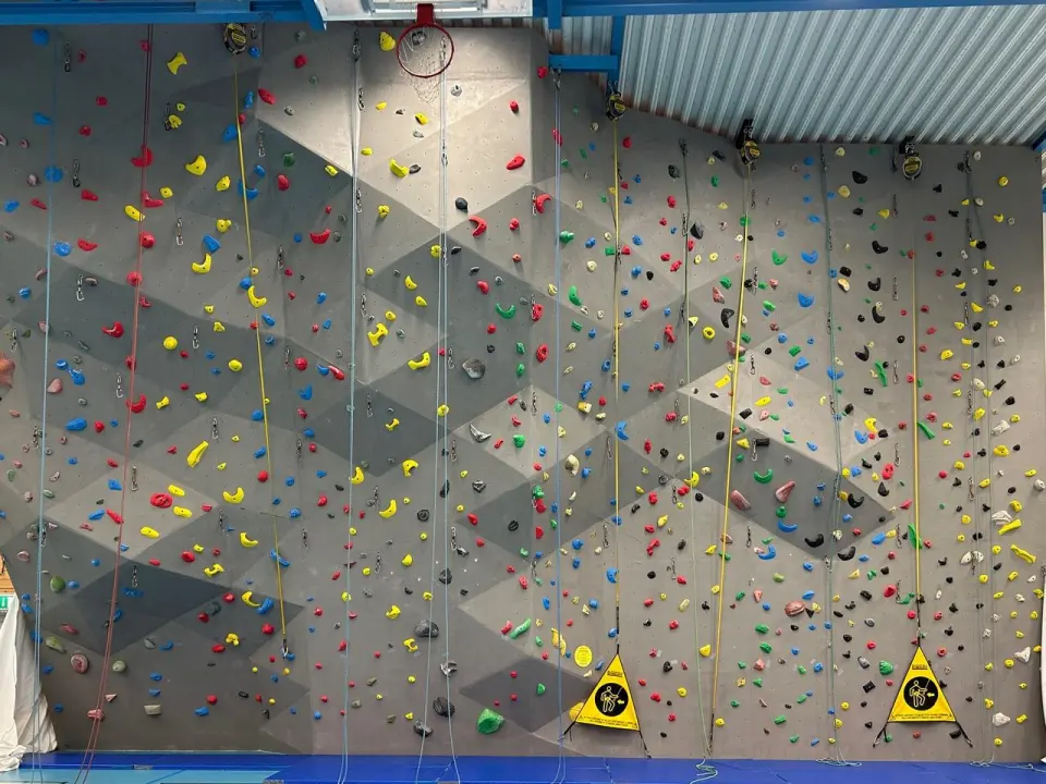 Grey climbing wall with coloured holds, auto-belays and blue crash mats in a sports hall