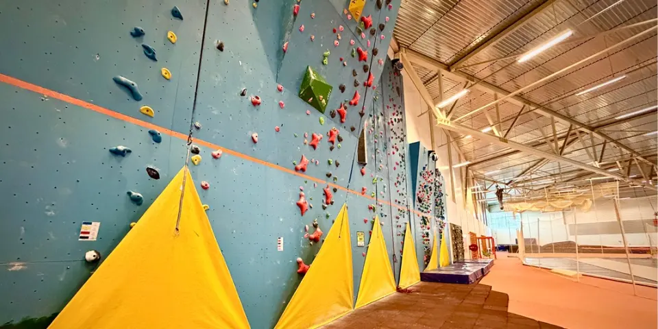Long blue climbing wall with yellow pads at its foot beside the arena's running track