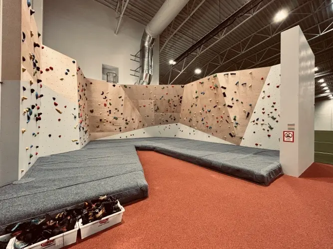 Bouldering wall of angled wooden and white panels above grey crash mats in a sports hall