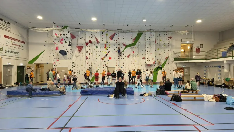 Roped climbing wall across the end of a sports hall, with climbers and belayers at its foot