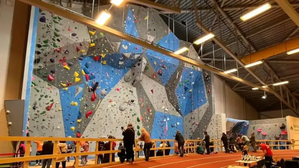 Tall climbing wall with grey and blue panels along one side of a sports hall, with climbers at its foot