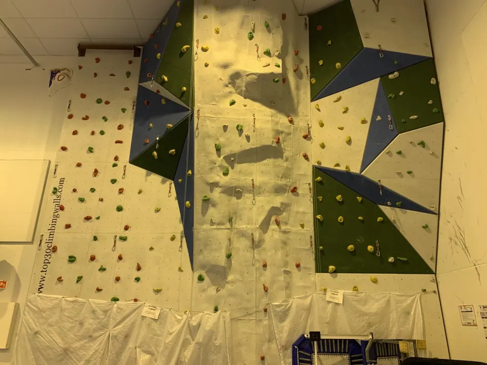 Climbing wall with green and blue angled panels and quickdraws in a gymnastics hall