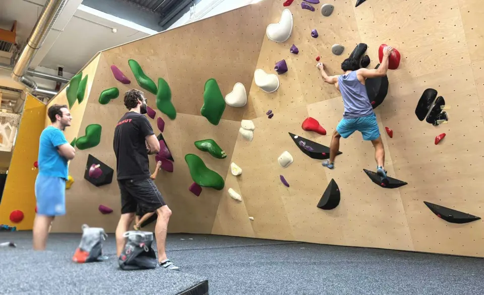 Climber on an angled wooden wall while others watch from the mats