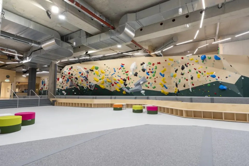 Long bouldering wall with colourful holds above grey mats and round seats