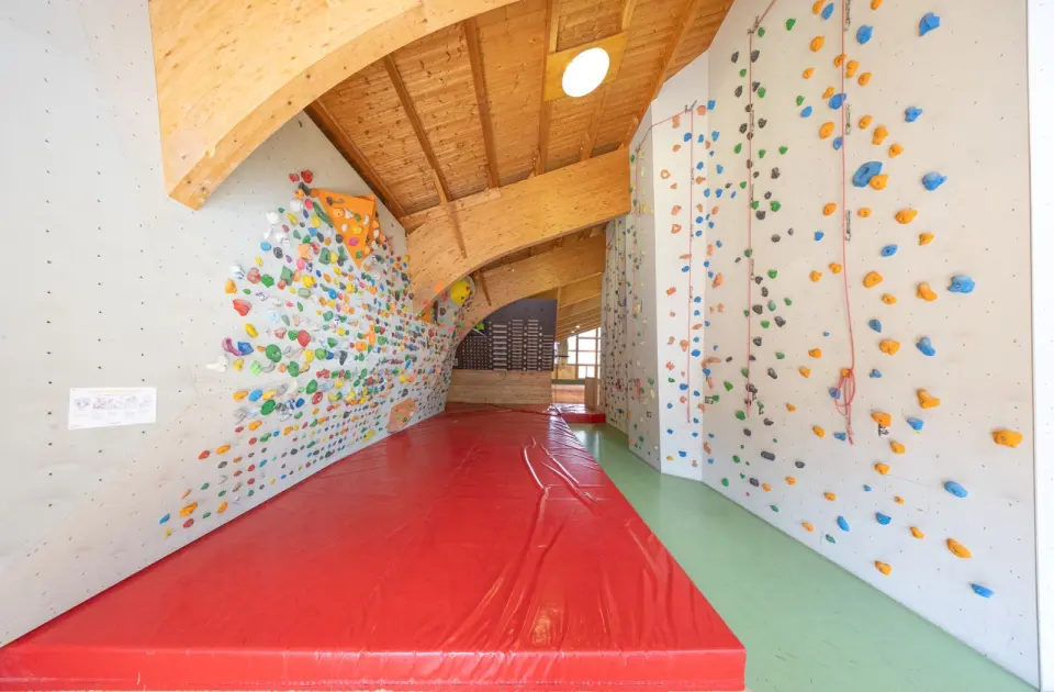 Bouldering wall over red mats and tall roped walls under a wooden roof