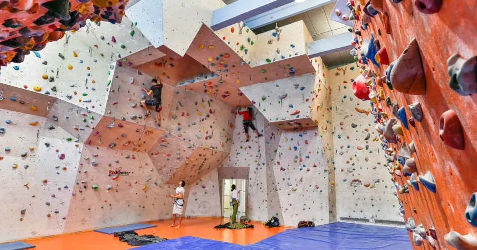 Two climbers leading on tall angular walls above orange flooring and blue mats