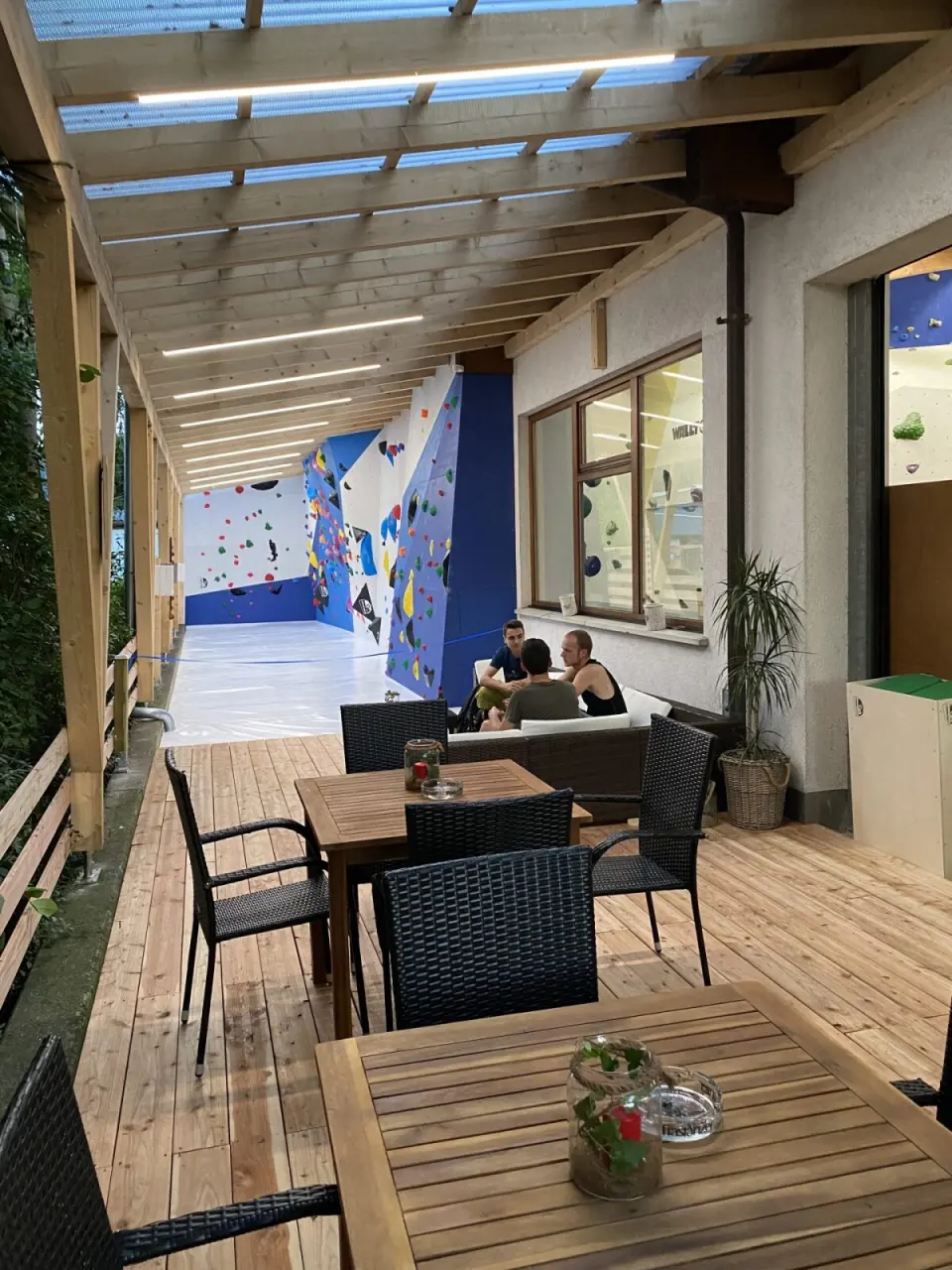 Covered wooden terrace with tables leading to the outdoor bouldering wall