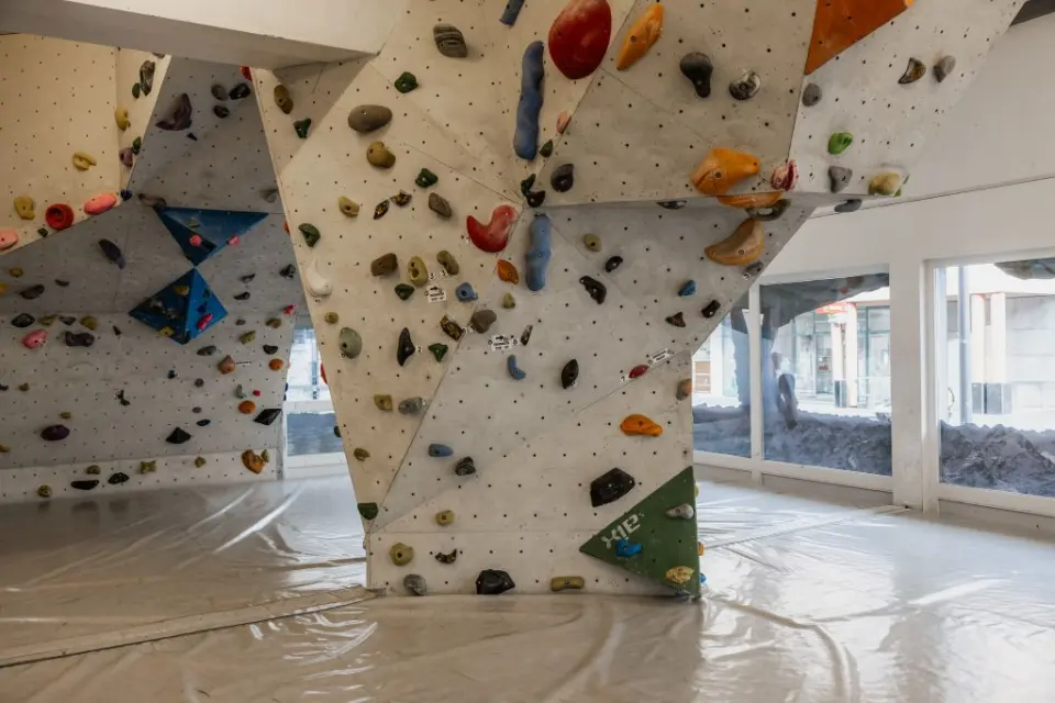 Angled bouldering pillars covered in holds above a matted floor, with windows behind