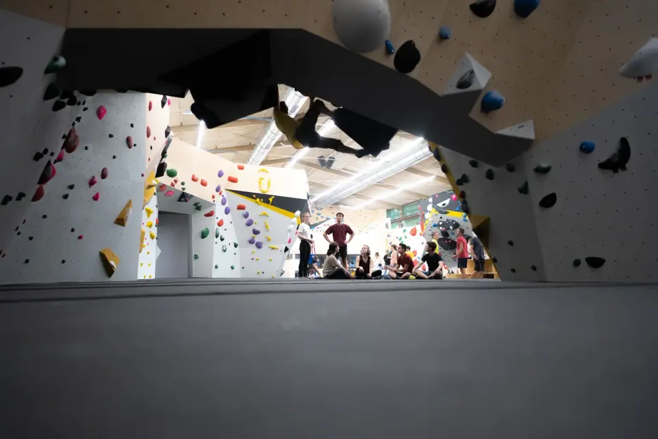 Bouldering hall seen through an archway in a steep wall, with climbers resting on the mats beyond
