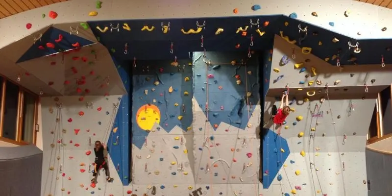 Roped climbing wall in a school gym with blue and grey panels, an overhang and two climbers