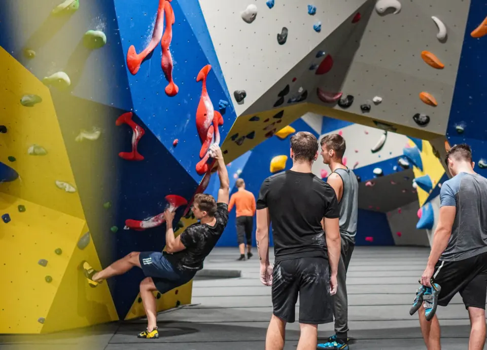 Boulderer swinging from red holds on a blue wall while others watch from the mats