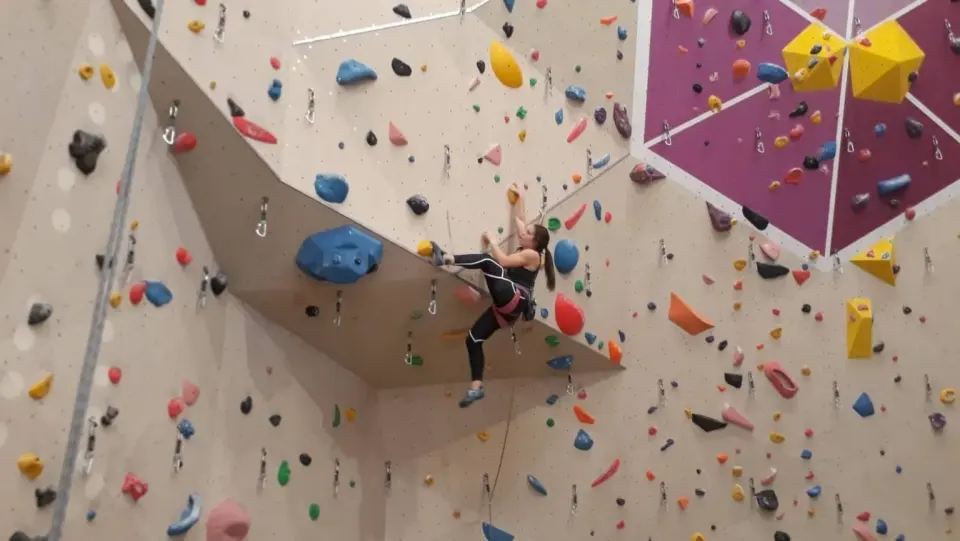 Climber on the steep lead wall at Boulderama in Klagenfurt, beside a purple panel with yellow volumes
