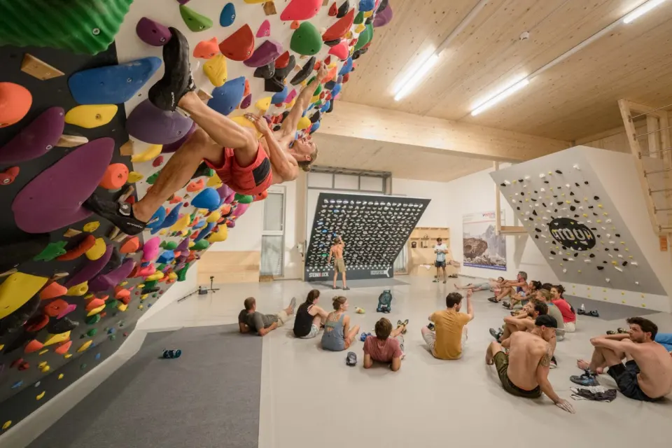 Climber on a steep wall packed with holds, with a training board in the background