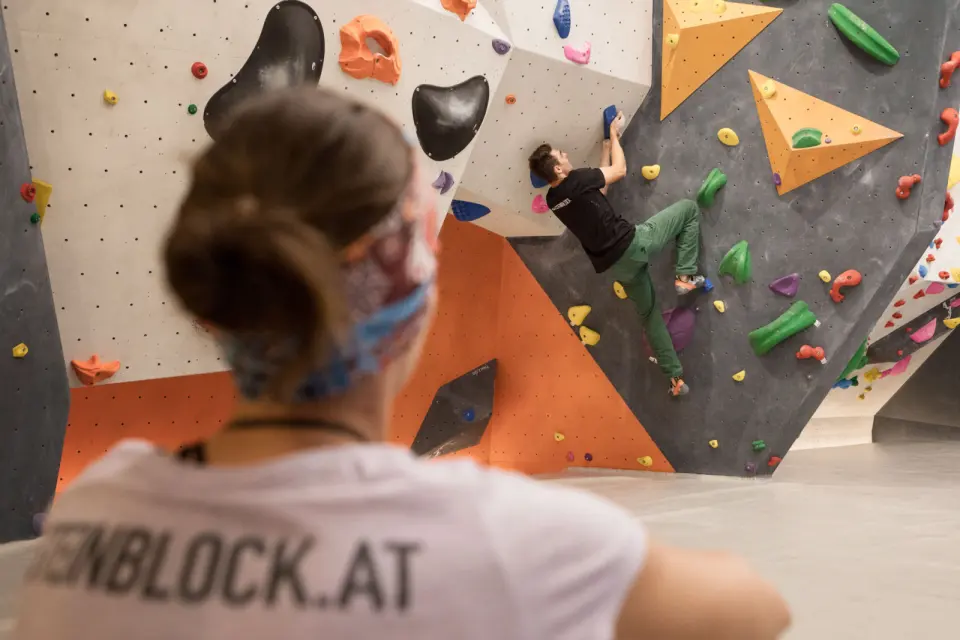 Climber on a grey wall with orange volumes, watched from the mats