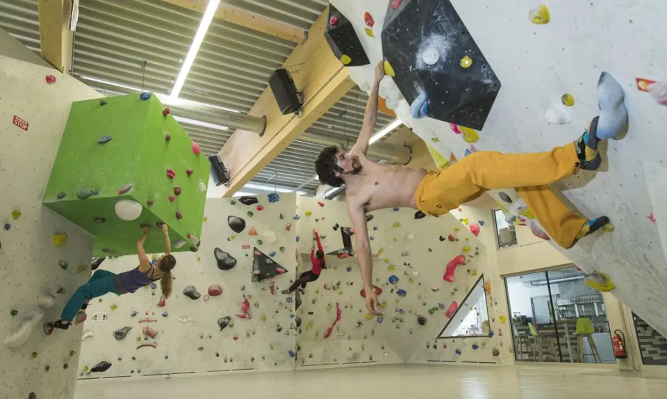Climbers on overhanging bouldering walls beside a large green cube-shaped volume