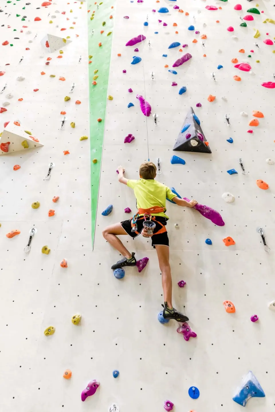 Young climber on a tall wall covered in colourful holds, seen from below