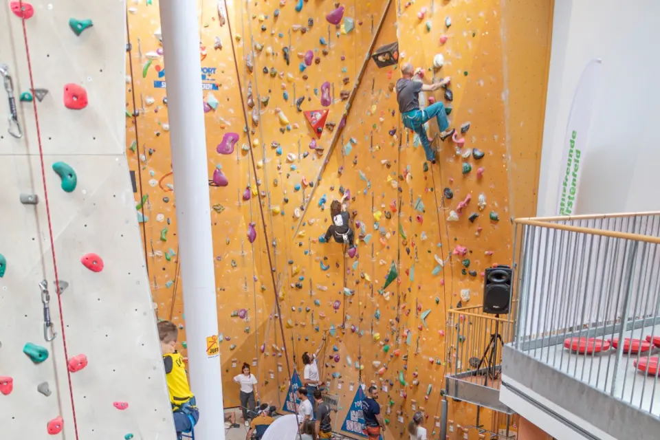 Two climbers high on the tall orange lead wall, with a grey climbing tower and a viewing gallery either side