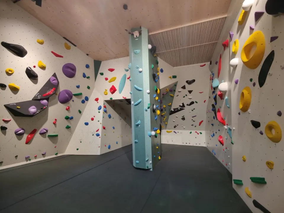 Bouldering room with a freestanding pillar, angled walls with coloured holds and a dark padded floor